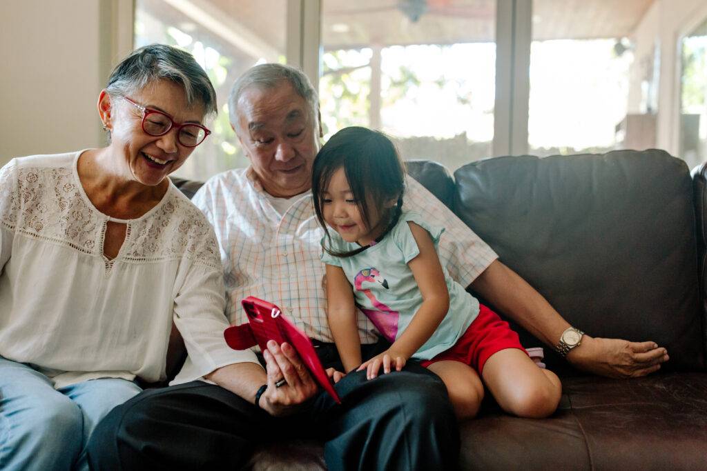 Happy Asian grandmother and grandfather sitting on leather couch next to smiling young girl.. They are looking at a cellphone.