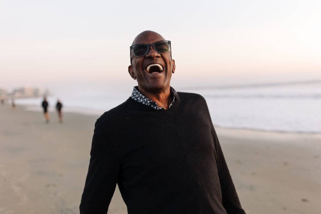 Portrait of joyful black man at the beach. He is smiling and wearing a sweater with checkered collared shirt.