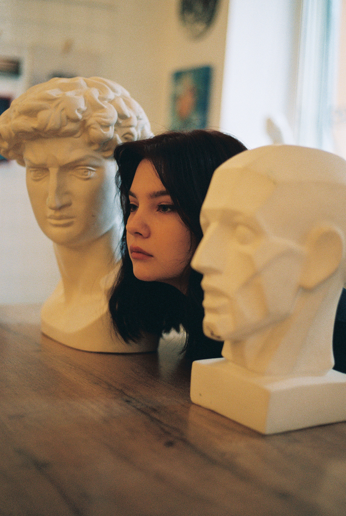 close-up film photo of a young woman's head in between white ceramic bust head sculptures in an art studio