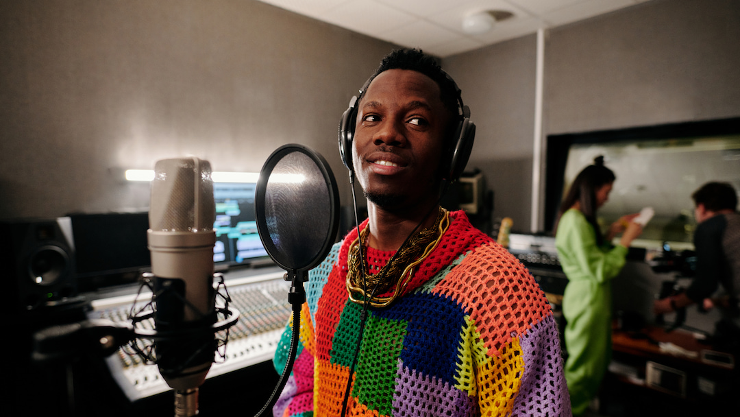 Handsome young Black man wearing stylish colorful outfit and headphones standing in front of microphone in recording studio