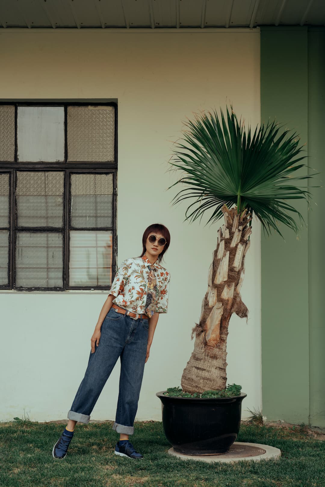 A young fashionable woman, exuding coolness, wears sunglasses, a tie, jeans, and sneakers, striking a playful pose imitating a tree beside a palm tree, while confidently looking at the camera. Her unique blend of style, fun, and confidence, creating an engaging and energetic visual representation.