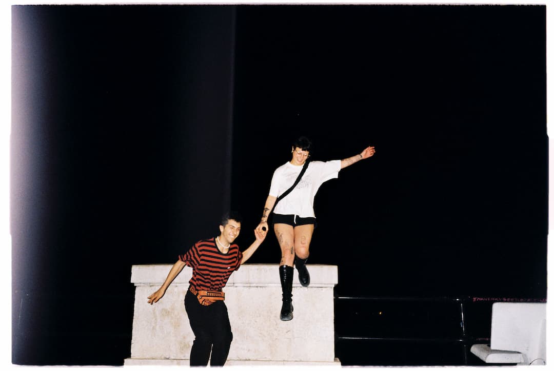 Boy and girl wearing punk-style clothes, boots and accessories. They are holding hands and jumping from a concrete structure in the city.
