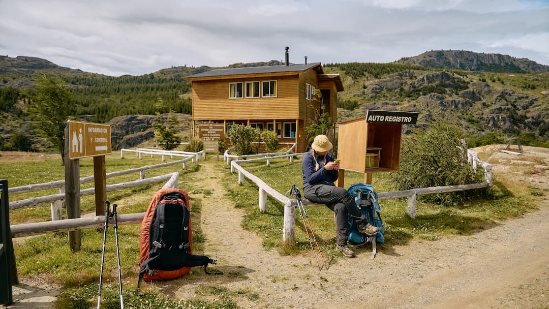 A woman is sitting in front of a wooden cabin at the start of a hike looking at her phone with her bag pack on the floor