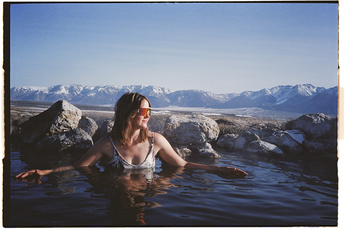 A young woman wearing sunglasses and bikini is enjoying a natural hot spring in the morning. She is facing the sun with her arms out feeling the warmth. Shot on 35mm Olympus XA3 film camera. Natural film grain, no added grain. Side view. Wild Willy's Hot Spring near Mammoth Lakes, California