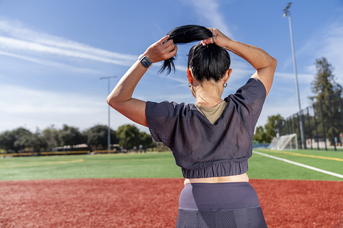 Athlete tying hair up in ponytail Back view of sporty athletic woman with dark black hair reaching her arms up tying her hair into a ponytail while standing on a soccer and baseball field looking away. She is preparing for physical exercise. Wearing black mesh top and skort. Photographed at a sports field with green and red turf surrounded by trees and blue sky above on a sunny day in California.
