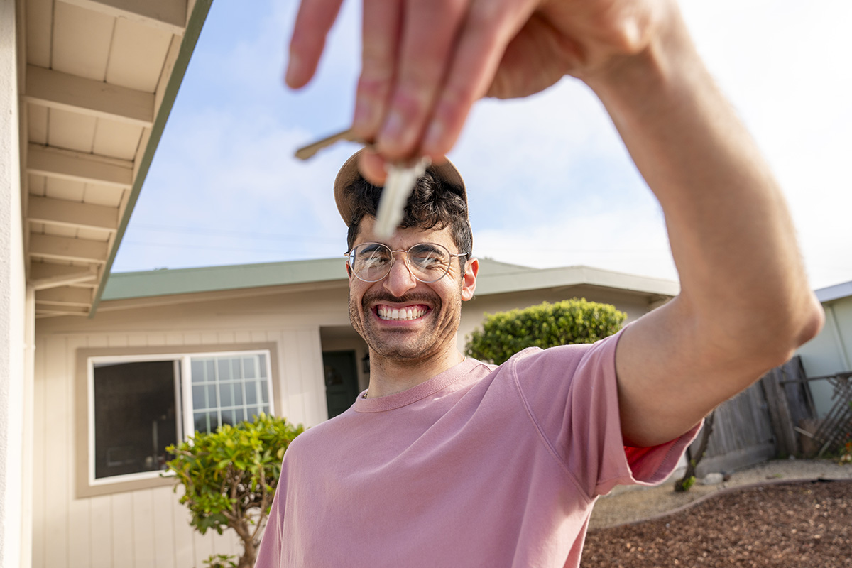 Excited new homeowner Portrait of young man smiling excitedly holding keys to new home standing in front of house frontyard, Milestone purchase