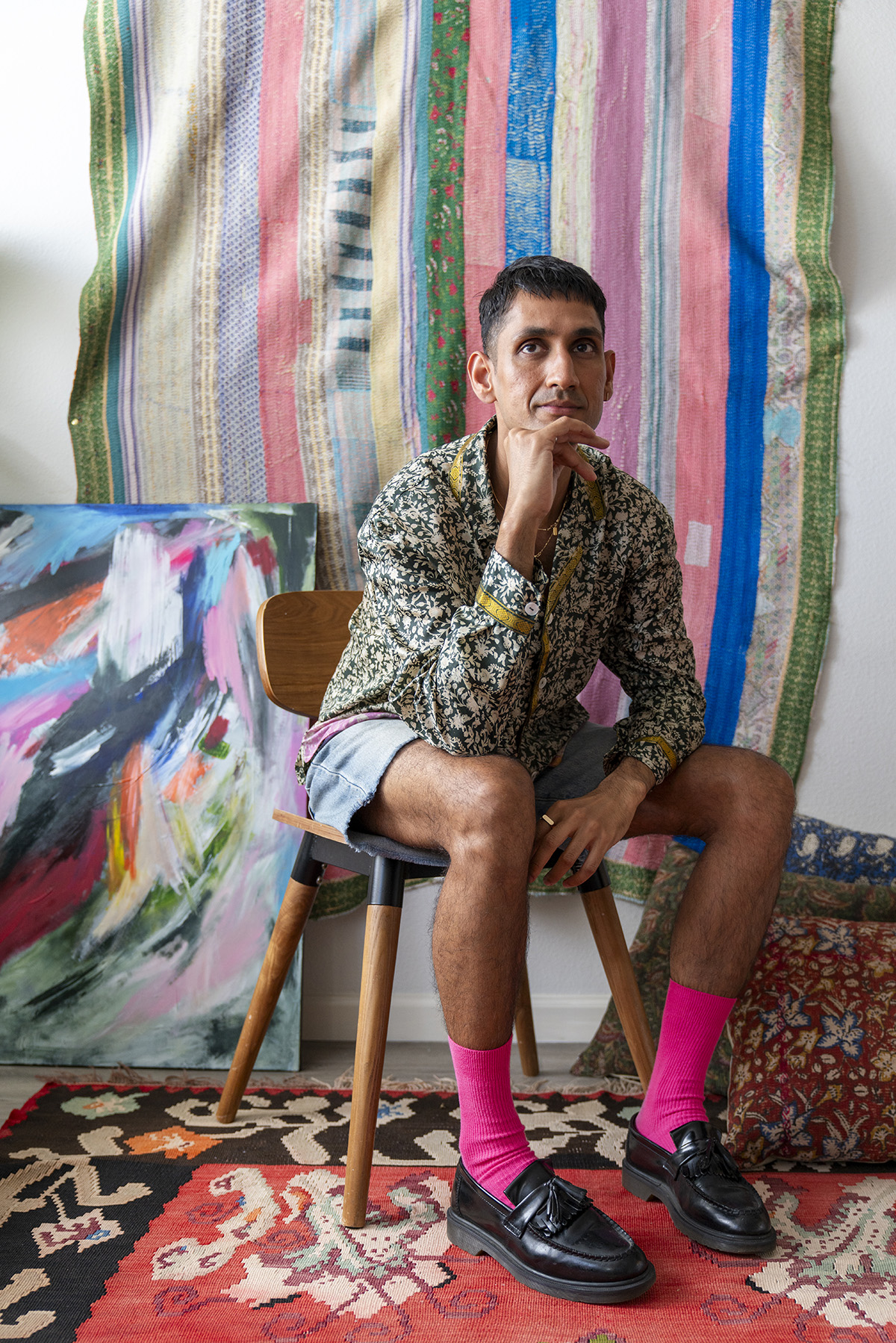 Fashion Portrait of stylish man Portrait of indian man at home with colorful backdrop sitting on wooden chair in colorful outfit and vibrant art in the background. Looking at camera with serious confident model like expression. Pink socks, denim shorts, black leather shoes.