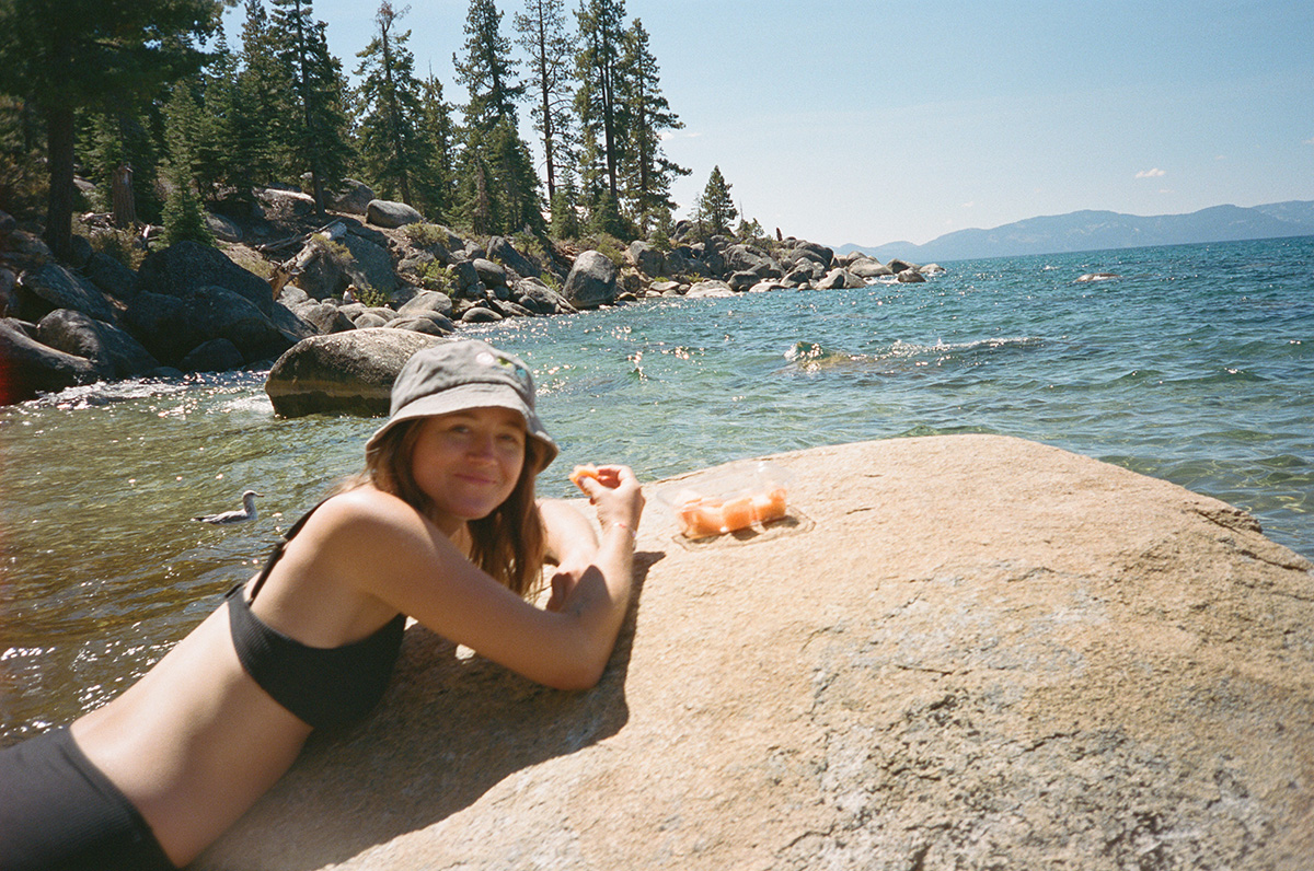 enjoying fruit snacking and eating laying on rock by lake surrounded by trees sunny day bikini and sun hat kodak portra 400 film stock camera aware authentic moment of woman soaking up sunshine on last days of summer vacation