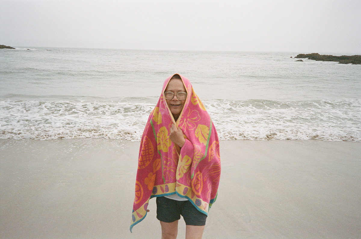 Retired older man standing on beach wrapped in tropical floral pink towel wearing eyeglasses and swim shorts cold from taking a dip in the pacific ocean beach waves overcast grey skies. Kodak Portra 400 film stock shot on olympus stylus. cold plunge