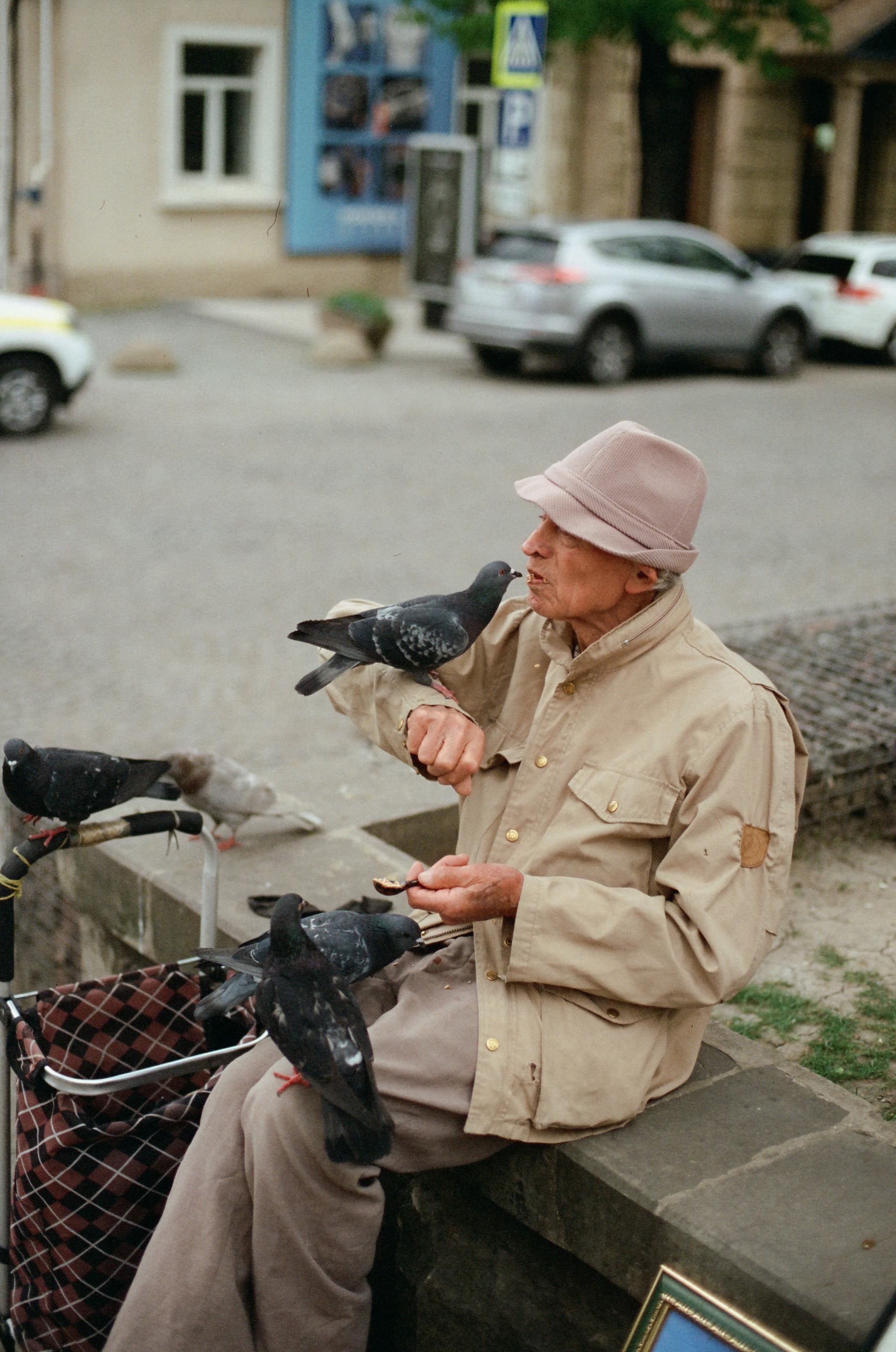 an elderly man in a beige jacket and pink hat feeds pigeons with grain from his mouth