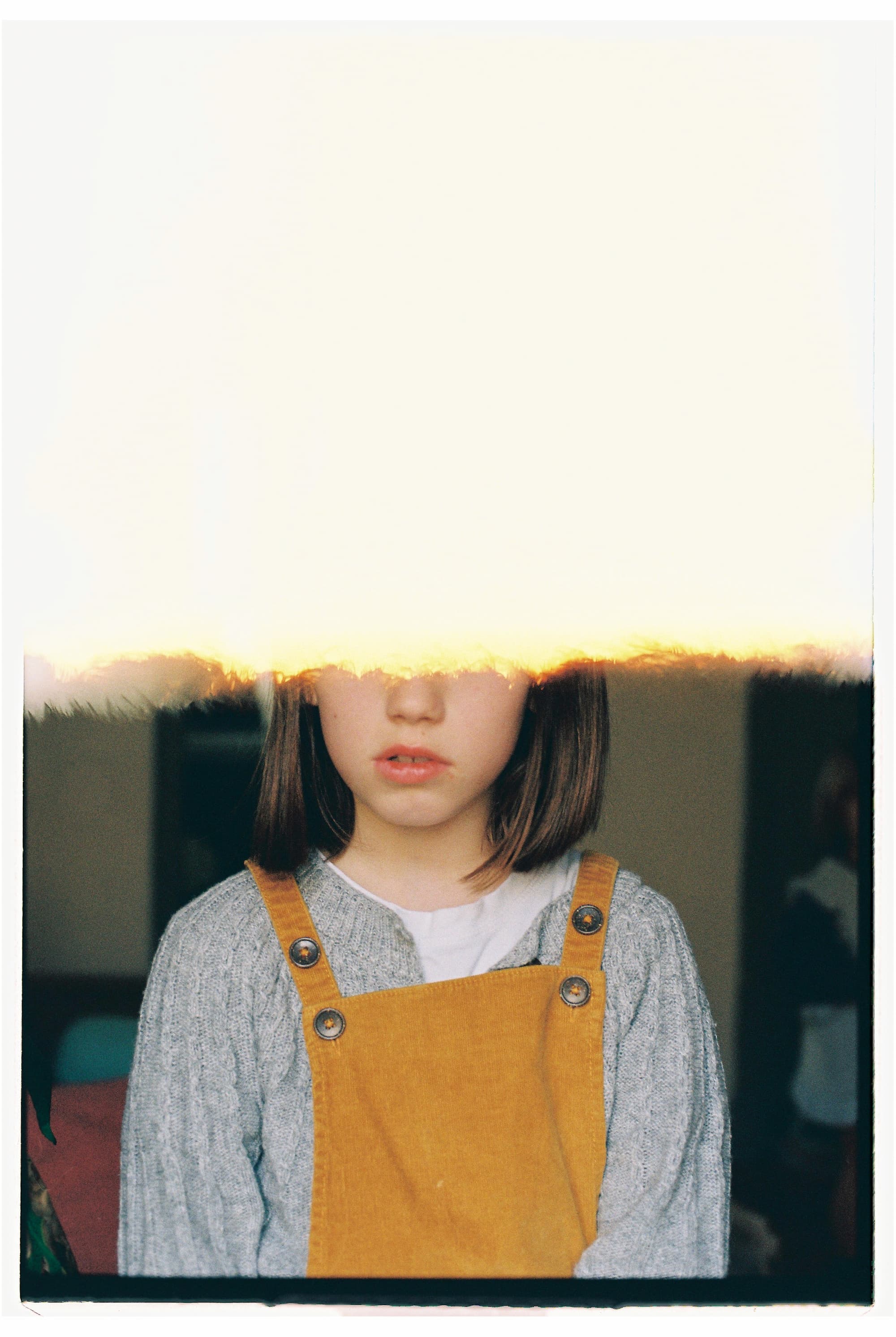 Part of a portrait of a girl looking at the camera with dark hair wearing a yellow corduroy dress and gray sweater, sitting on a couch at home.