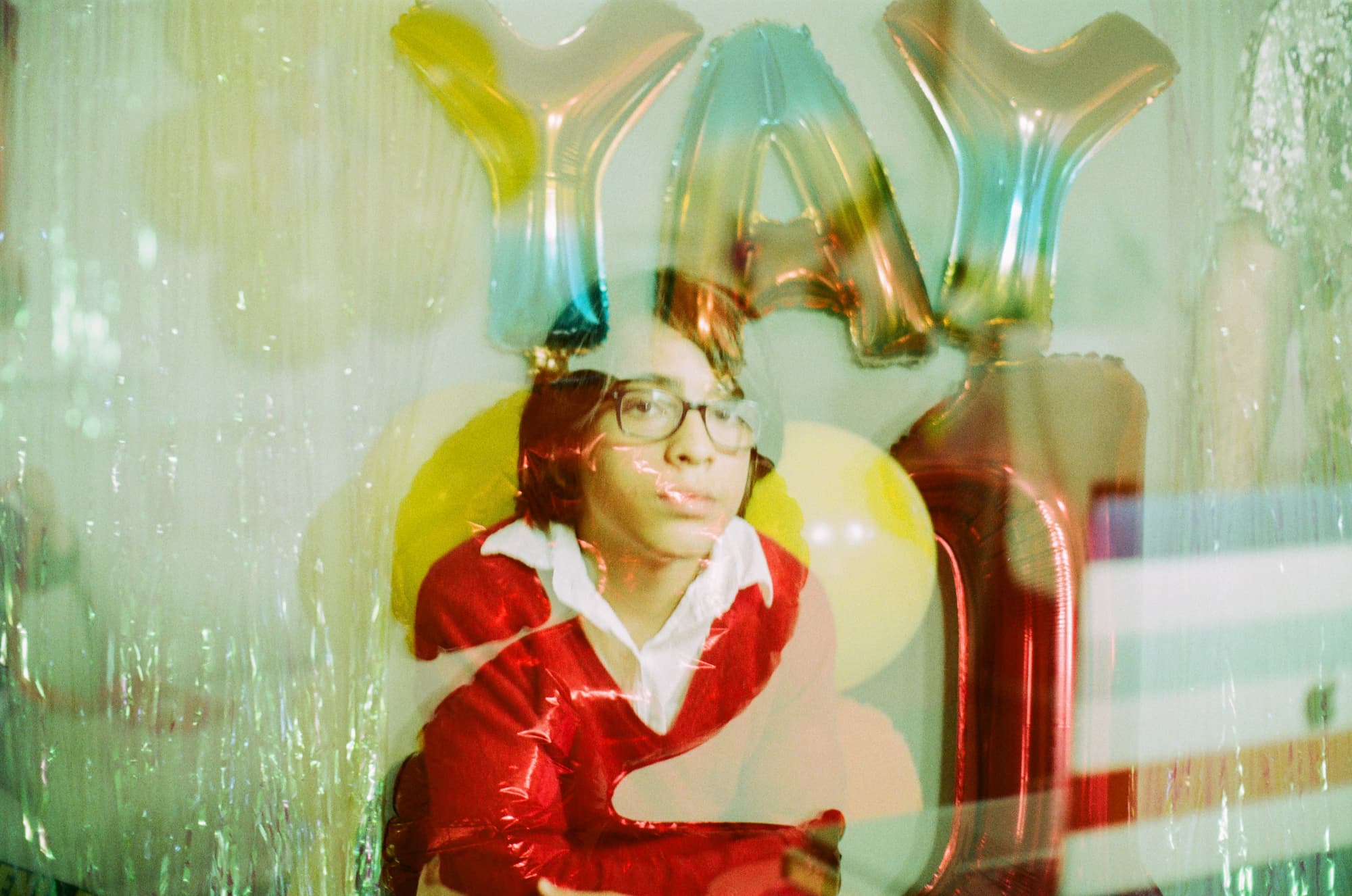 Tween boy looking at the camera through a reflective surface with colorful balloons saying YAY in the back. 35mm analog film and double exposure