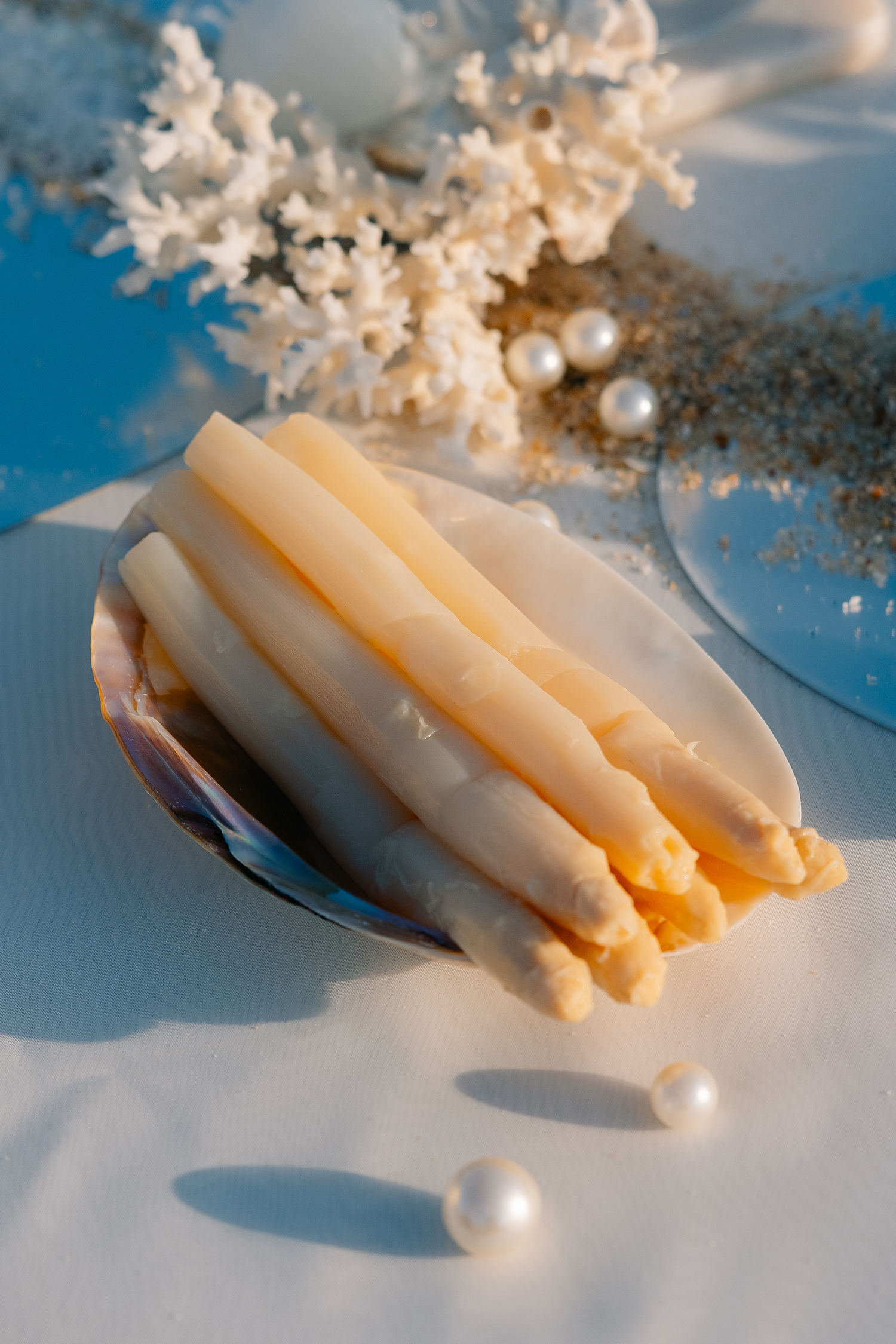 White Asparagus on Shell with Coastal Decor A close-up of white asparagus spears displayed on a shell, surrounded by coral, pearls, and sandy elements, evoking a serene coastal ambiance