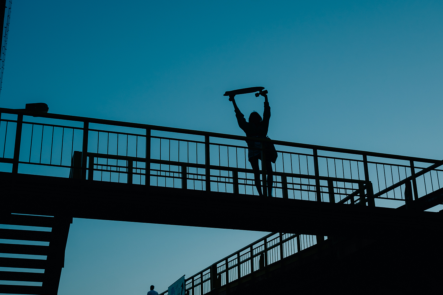 Silhouette of a person holding a skateboard on a bridge at sunset Silhouette of a person raising a skateboard on a bridge during sunset, capturing a moment of youthful freedom.