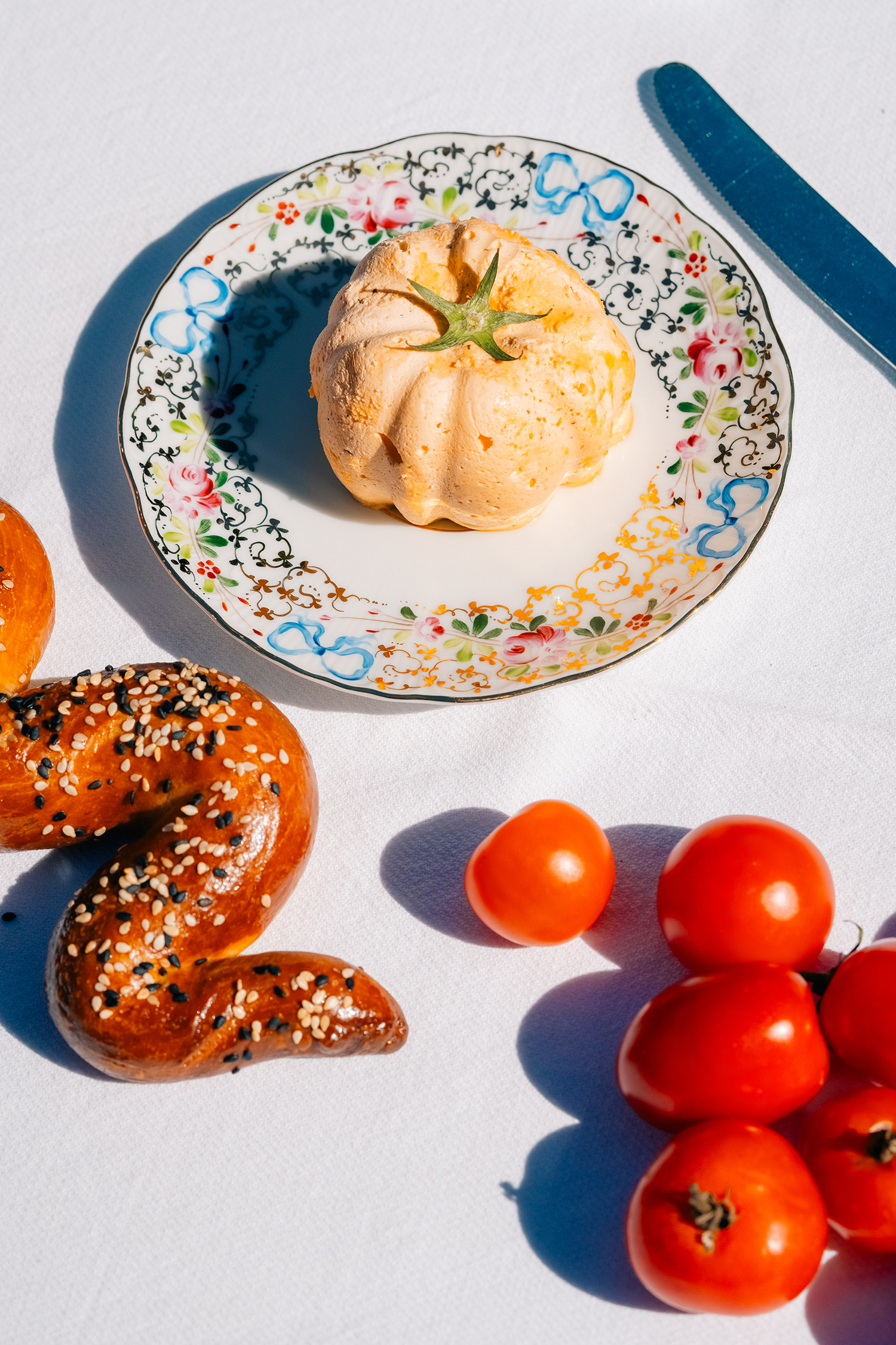 Colorful food arrangement with pastry, tomatoes, and a bread twist A vibrant table setting featuring a pastry on a decorative plate, fresh cherry tomatoes, and a sesame-topped bread twist.