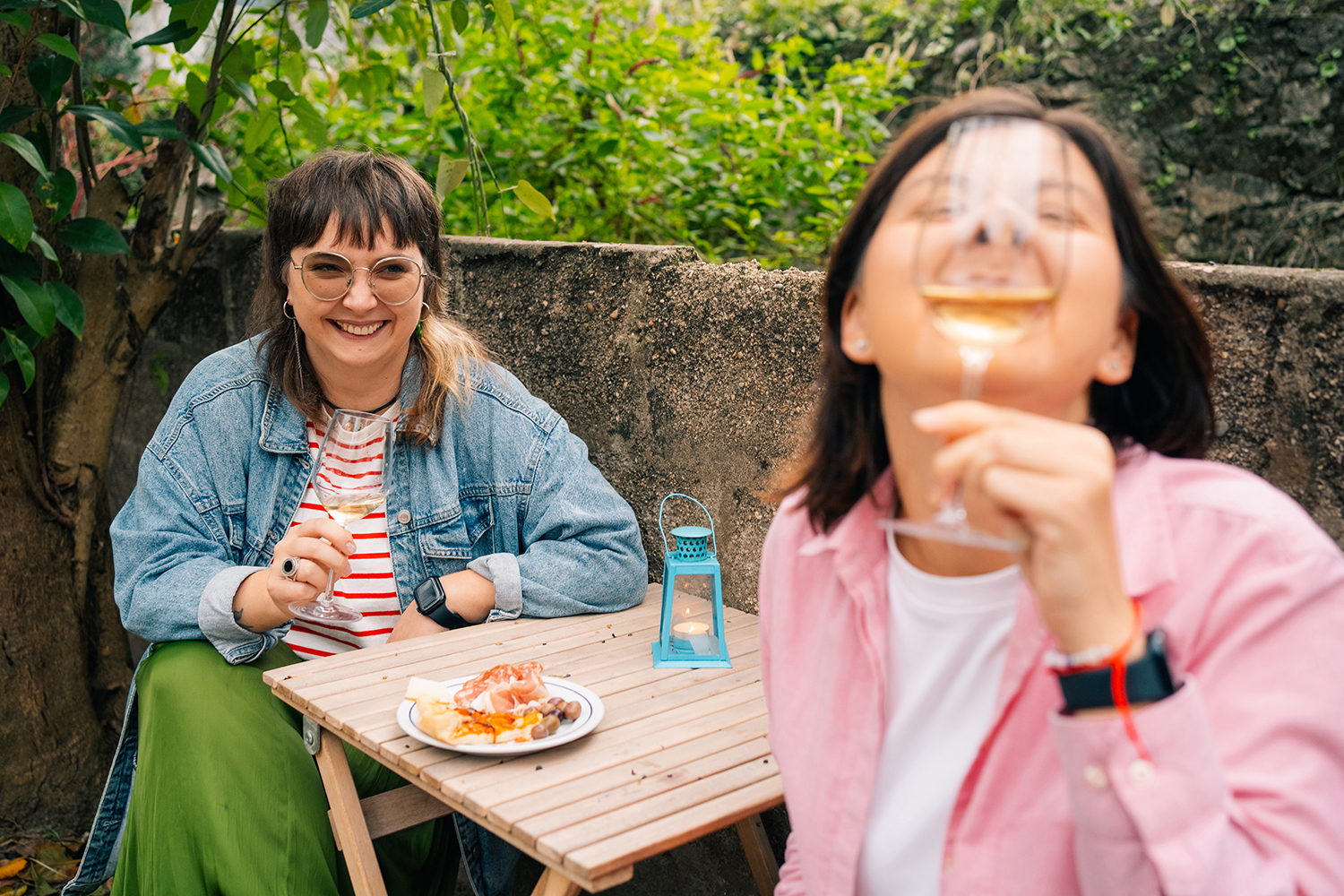 Friends Enjoying Drinks and Laughter Outdoors Two women sharing laughter and drinks at an outdoor setting, with a rustic table and food platter.