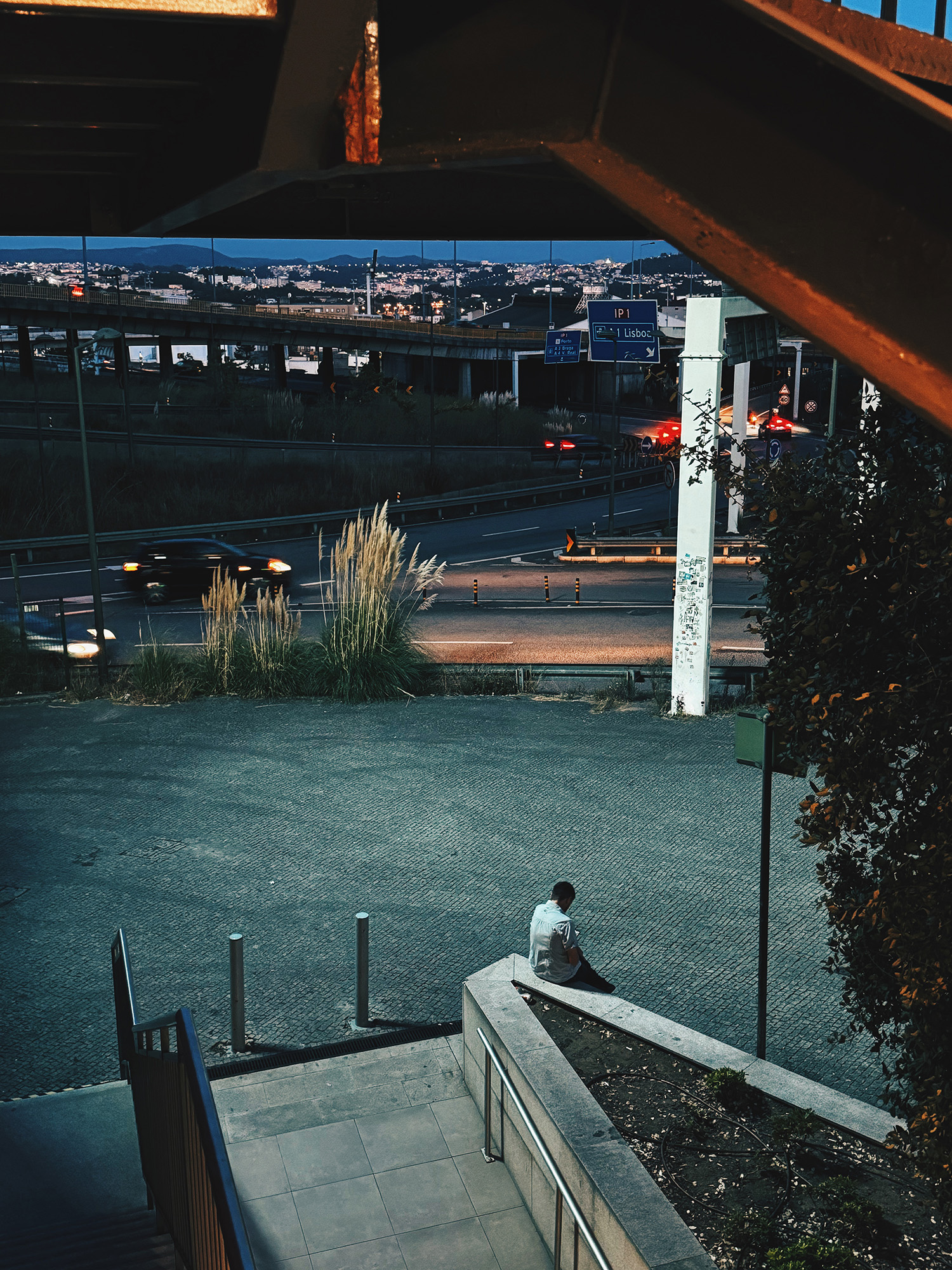 Urban Scene with Person at Twilight A person sitting on a stairwell in an urban setting, with traffic lights and cityscape in the distance.