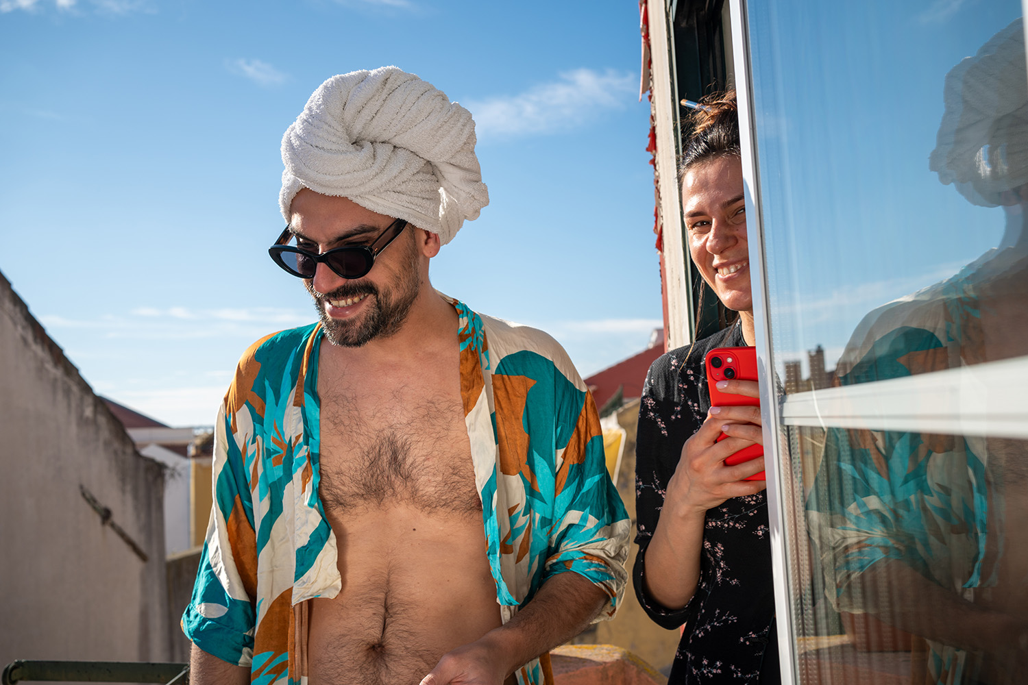 Couple Enjoying Relaxation Outdoors A man in a colorful shirt and towel turban smiling with a woman holding a red phone, standing outdoors under sunlight.