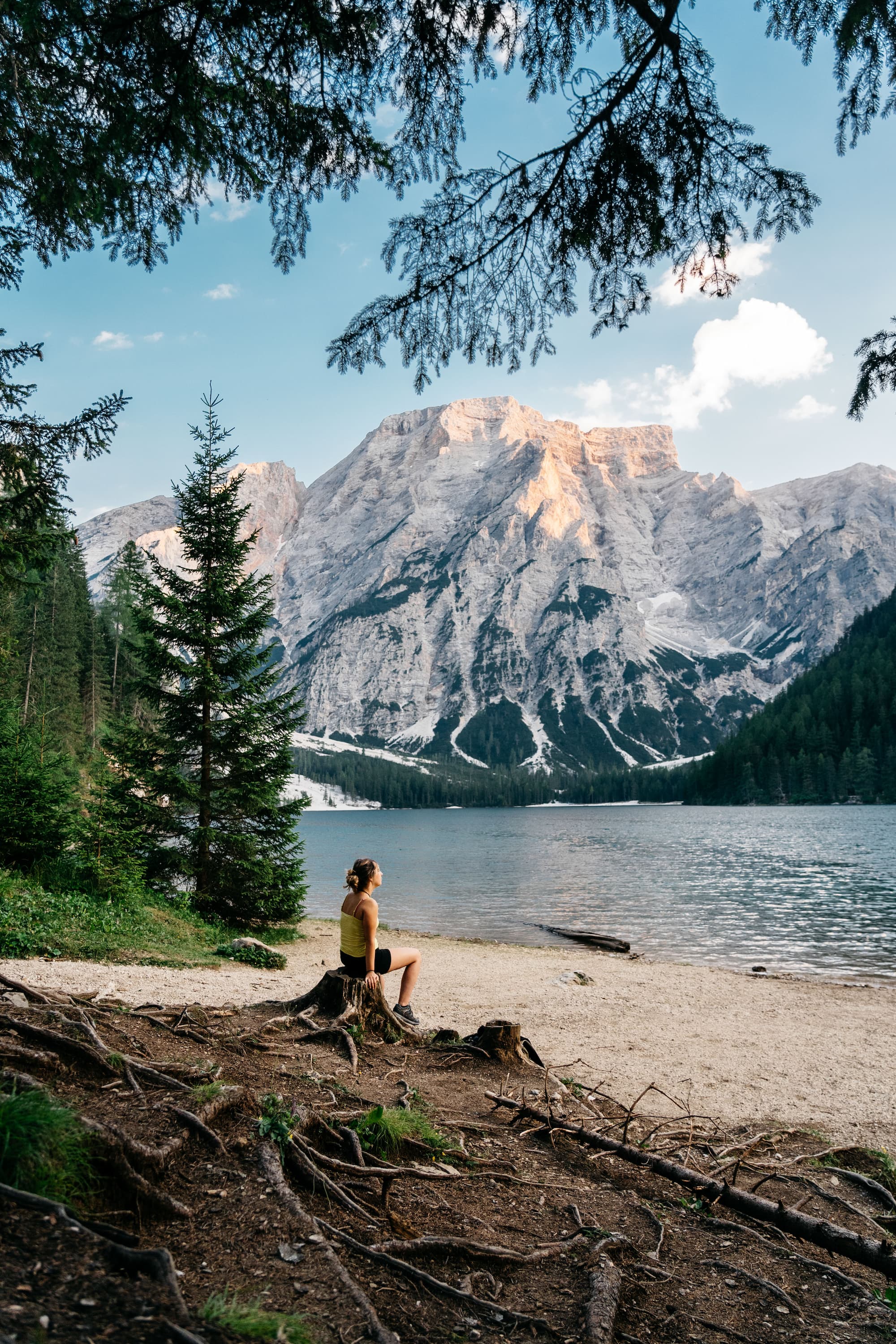 Side view of woman in sportswear sitting on stump at lake and contemplating beautiful landscape with snowy mountains in Lago di Braies, Italian Dolomites