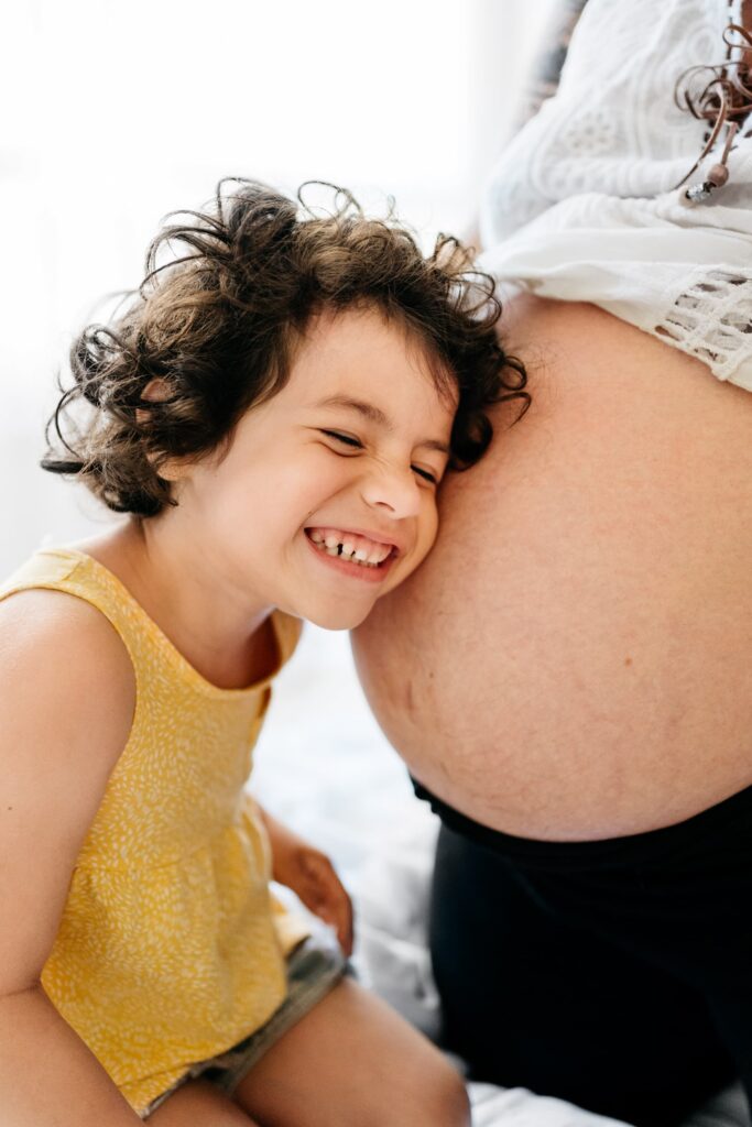 Happy mixed race girl smiling and listening to baby in pregnant tummy of crop mother at home