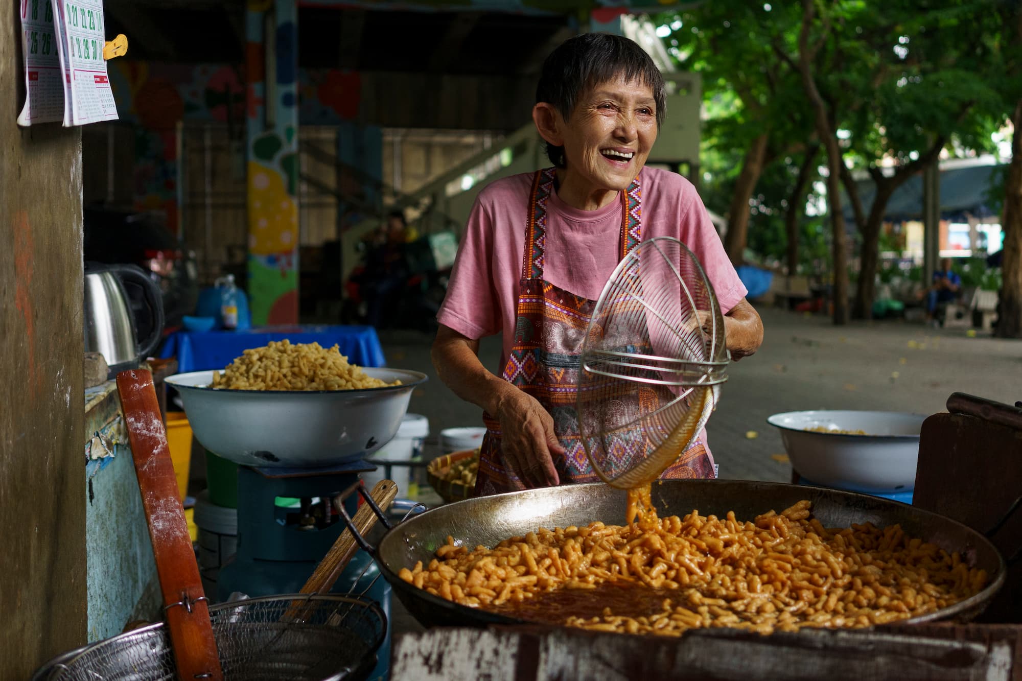 An elderly Southeast Asian woman smiles while cooking a crispy, mini version of Pa Tong Ko, a Thai appropriation of the Chinese-born fried dough stick Youtiao, from a large wok filled with hot oil at a local street food vendor in Talat Phlu, Bangkok, Thailand.