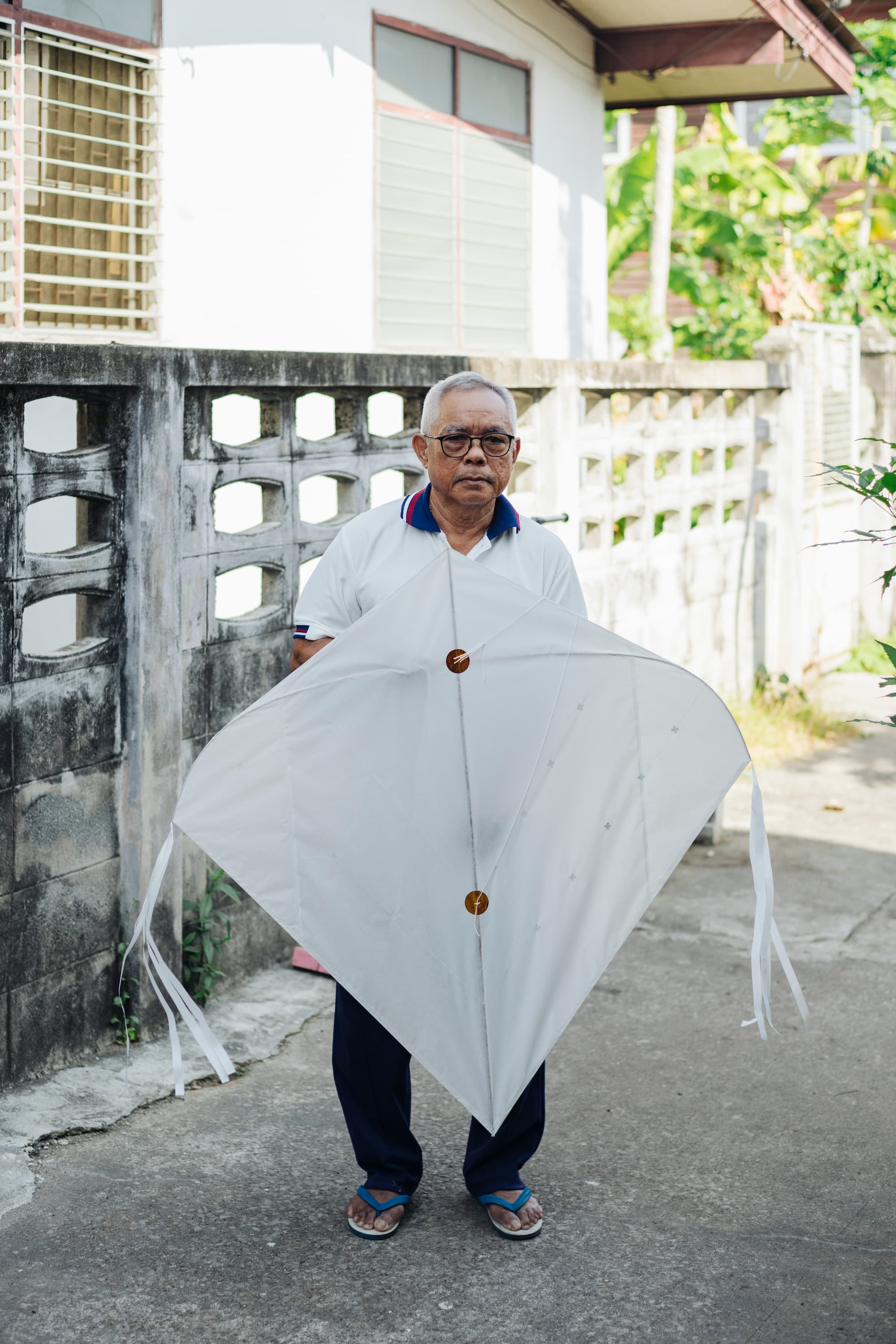 Portrait of an elderly Southeast Asian man holding an E-Lum kite, a traditional Thai kite, with his hands outside his kite-making workshop in Bangkok, Thailand.