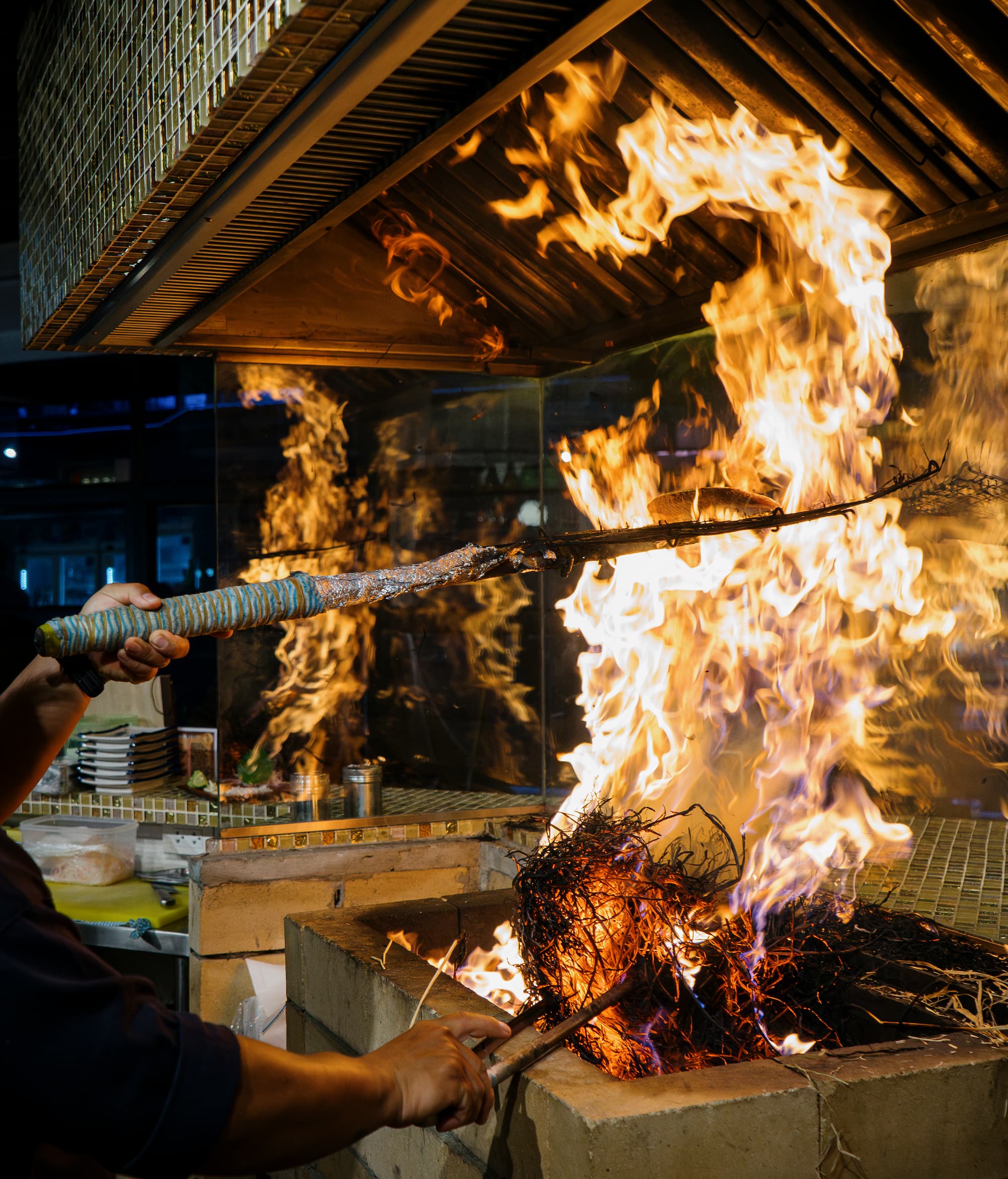 A chef grilling large piece of fish meat using Warayaki, a traditional Japanese technique that burns straw to add a smoky flavor to quickly cooked food, at the open kitchen inside a Japanese restaurant in Bangkok, Thailand, at night.