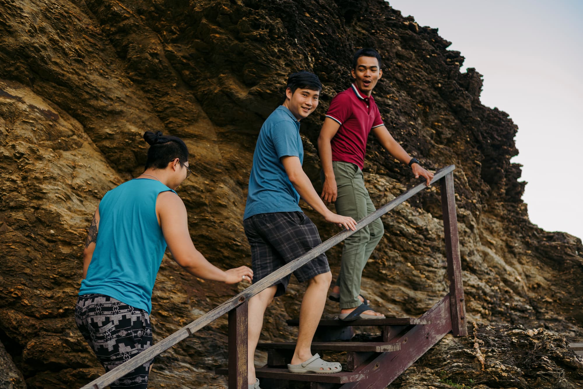 A group of young Southeast Asian men is enjoying their vacation while ascending a set of wooden stairs near a rocky coastline at sunset on the island.