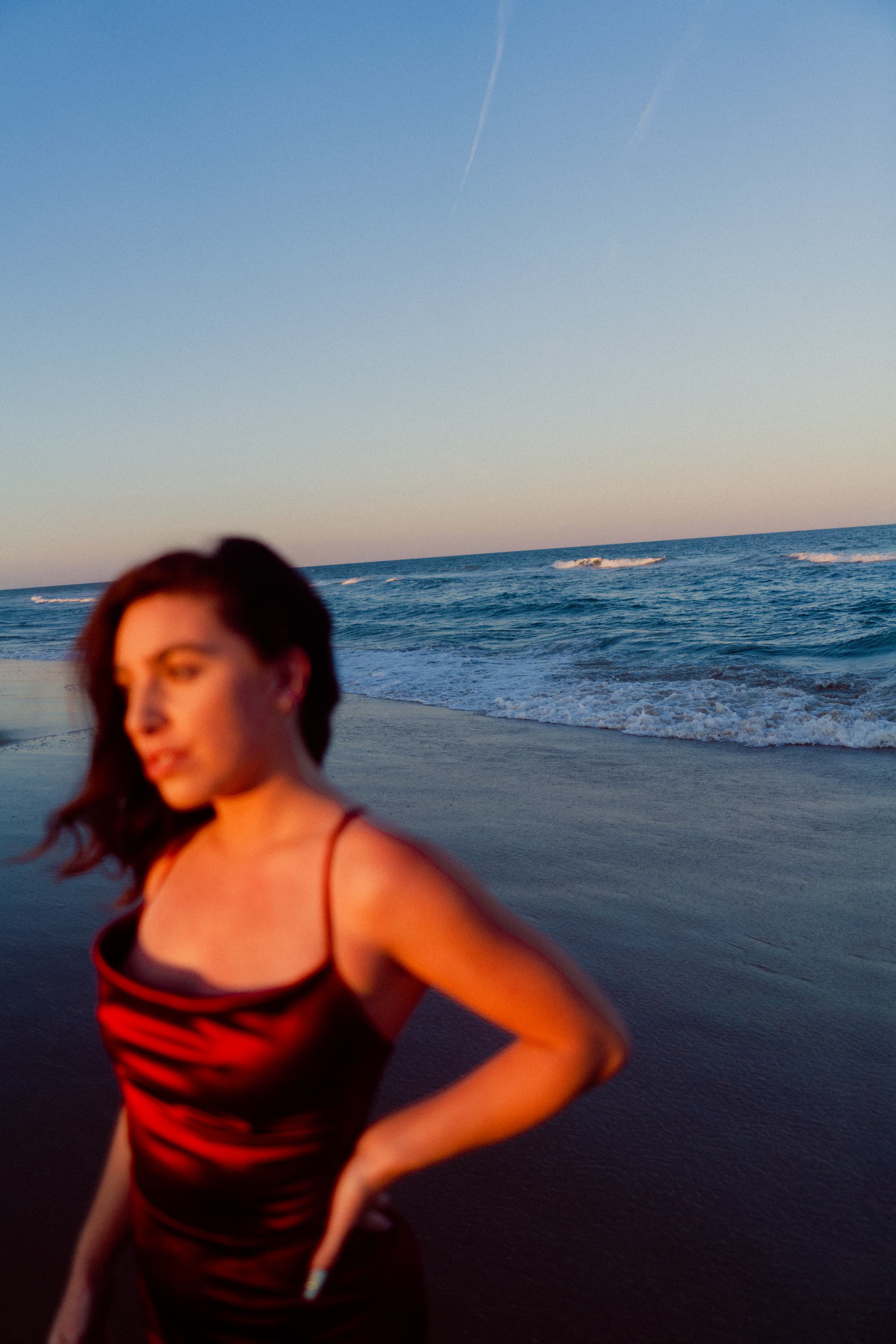 A beautiful young woman in a red dress standing at beach shore during high tide with waves in focus