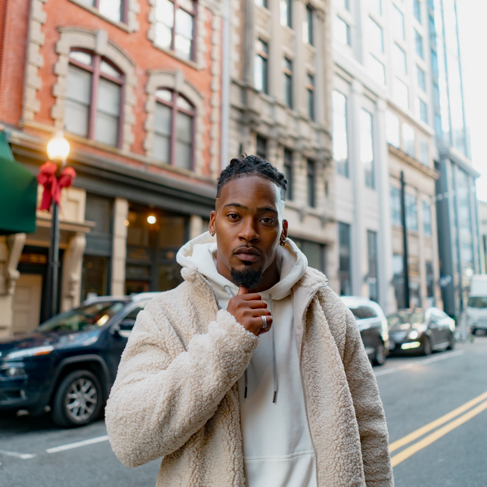 A close-up portrait of a young Black man posing in a downtown city street. His expression is poised and confident, with city architecture and street activity softly blurred in the background, highlighting his presence against the city’s dynamic energy.