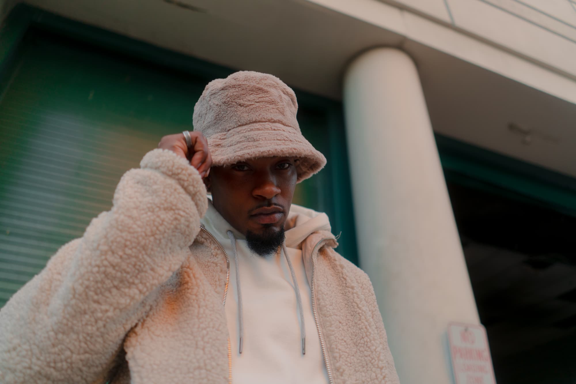 A low angle photo of a young black man standing near a building in a city, with his hand on his bucket hat.