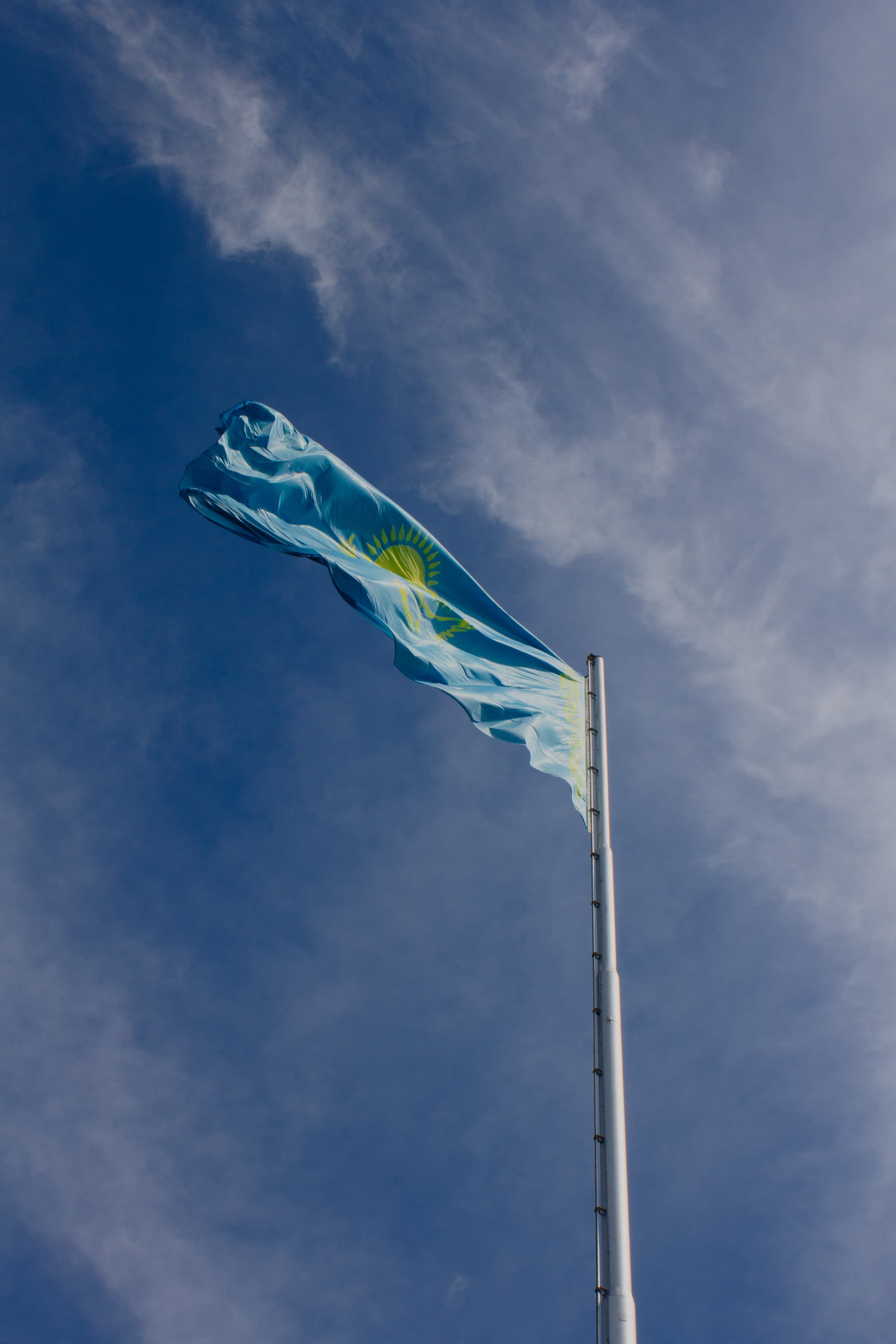 The vibrant blue and gold Kazakh flag ripples dynamically against a deep blue sky, its intricate sun and eagle design catching the sunlight. Each gust of wind reveals the flag's full glory, symbolizing national pride and the vastness of the steppe.