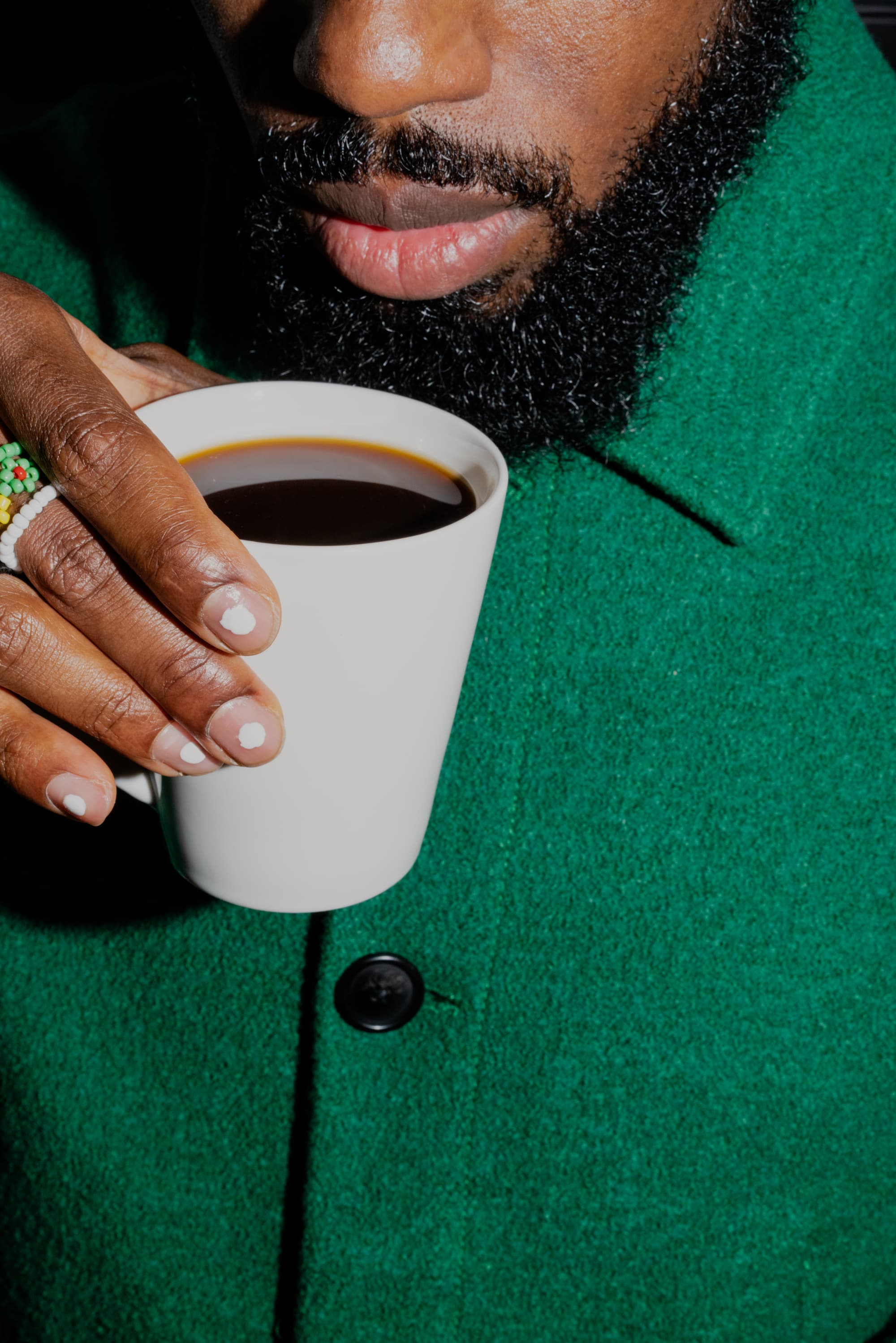 Young bearded black man raises is mug to take a sip of his coffee in green wool coat, with painted nails and beaded rings peeking into frame.