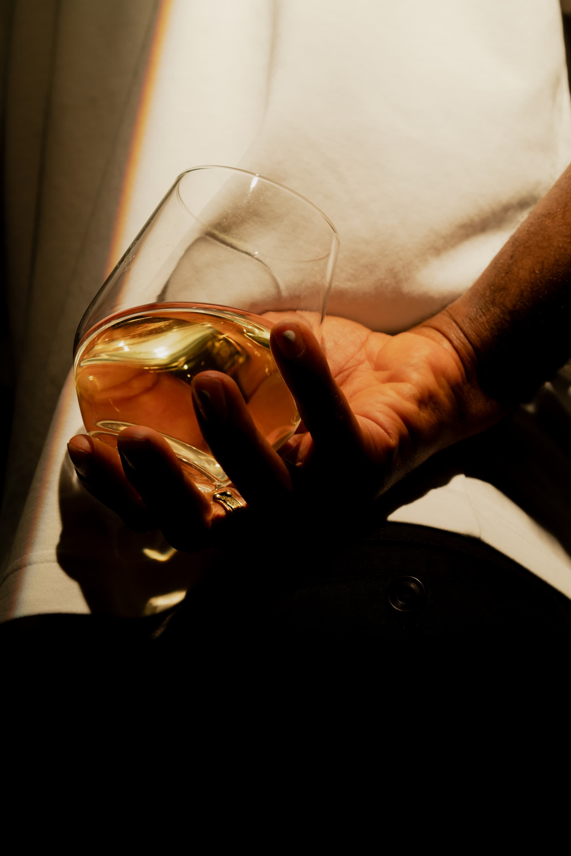 A striking portrait captures a man with a glass of white wine held discreetly behind his back, the overhead lighting casting dramatic shadows that accentuate his confident posture. The chilled glass gleams subtly in the low light, its condensation and pale hue contrasting with the moody atmosphere—hinting at sophistication and mystery.