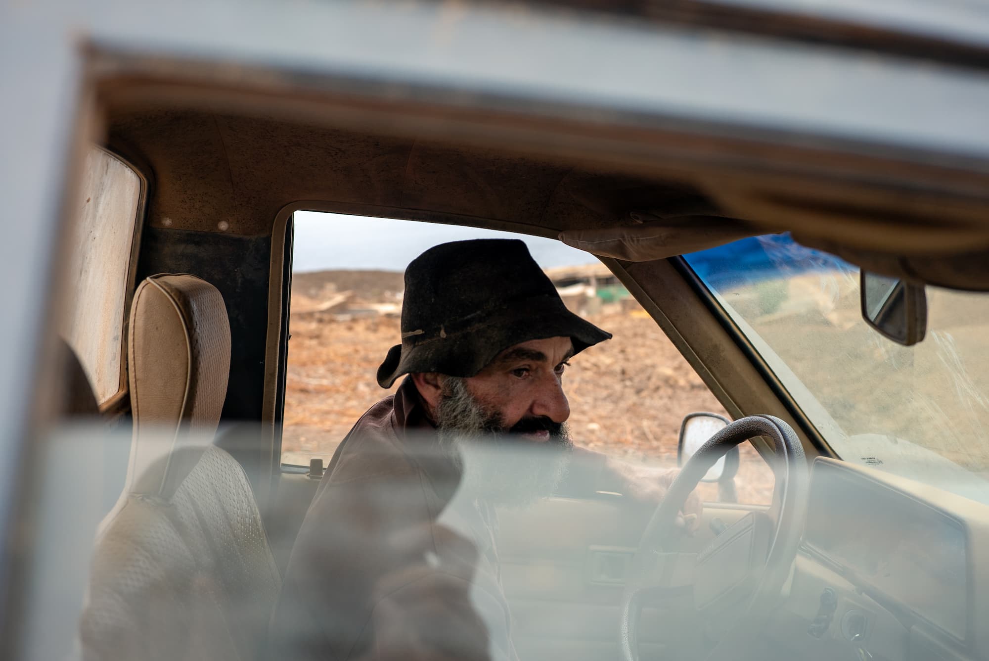Close-up portrait, through a window, of an older bearded person sitting in the driver's seat of a car during the daytime. An elderly man drives a pickup truck.