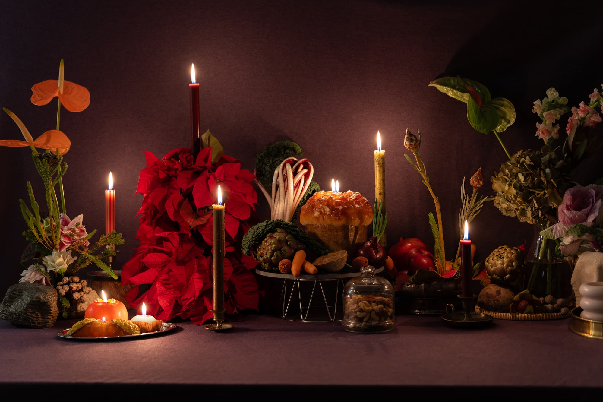 A lavish still life featuring a radiant poinsettia as the centerpiece, surrounded by an array of festive elements. The composition includes glowing candles, a persimmon candle, vibrant winter vegetables, nuts under a glass dome, and a panettone lit from within. Set against a textured purple backdrop, the arrangement evokes a sense of warmth, celebration, and the richness of the holiday season.