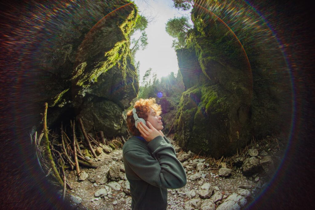 A happy red-haired guy enjoys music with headphones on a sunny day against a beautiful forest landscape. Portrait of a happy guy in the woods on a fisheye camera.