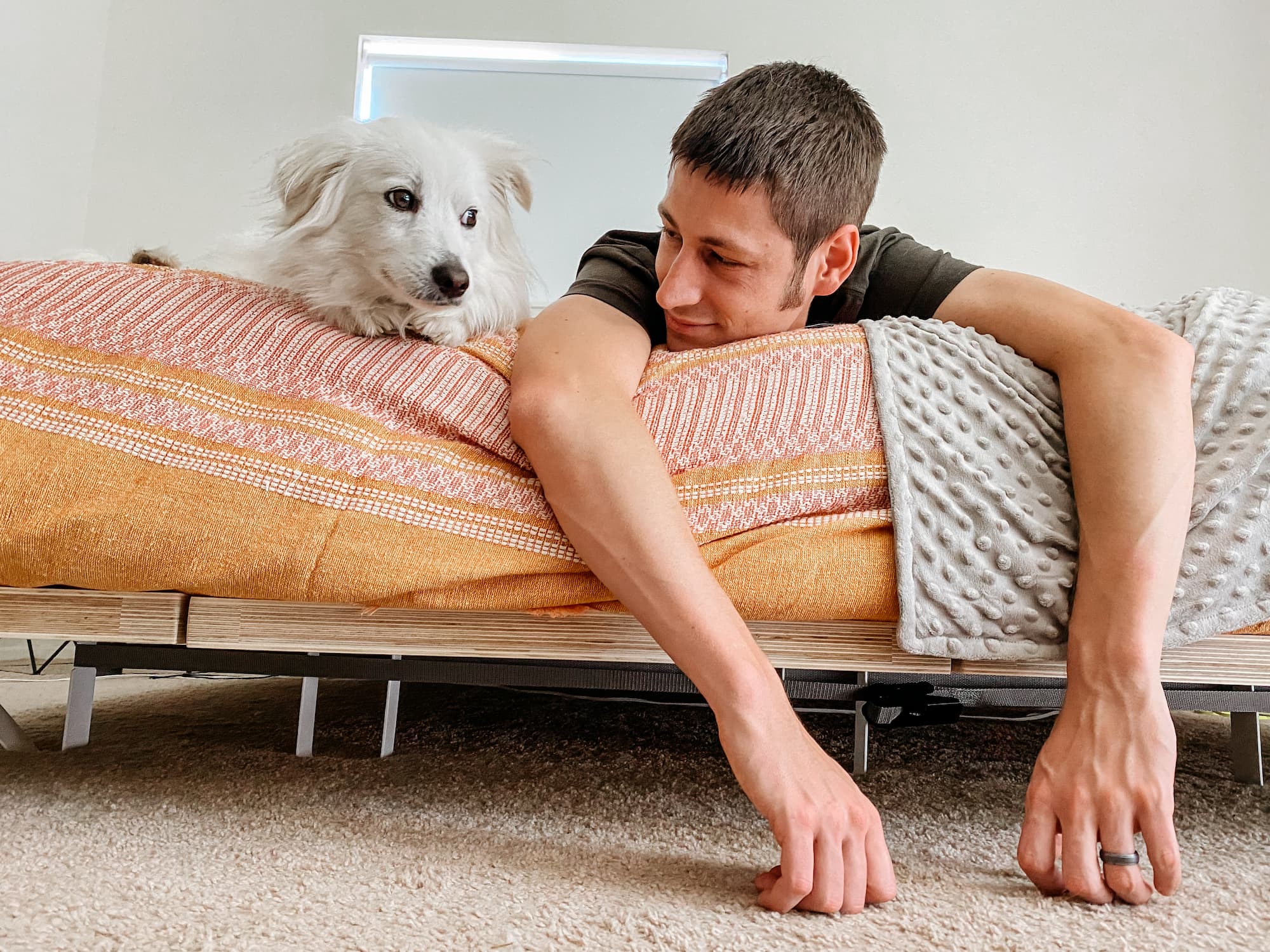 a man and a white fluffy dog companionably lay at the end of a bed while looking at each other. the bedroom has white walls and the bed is covered in an orange blanket. the man's arms hang over the edge of the bed.