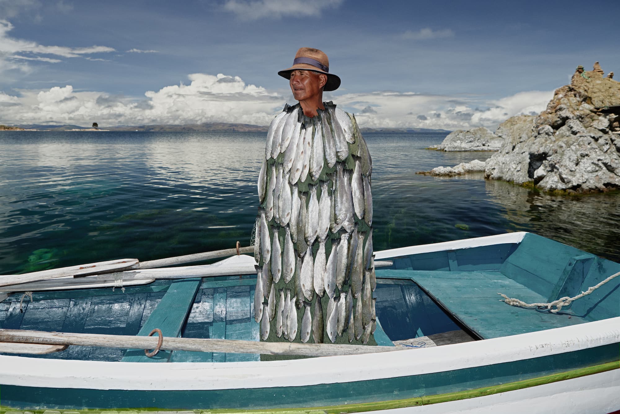 A Bolivian fisherman standing still on a wooden boat floating on Lake Titicaca, wearing a unique handmade poncho entirely covered in real trout. Conceptual and surreal outdoor portrait combining elements of traditional lifestyle and contemporary art. Shot in natural daylight with clear blue water, rugged shoreline, and dramatic clouds in the background. The image blends documentary and fine art styles, representing identity, culture, and symbolism.