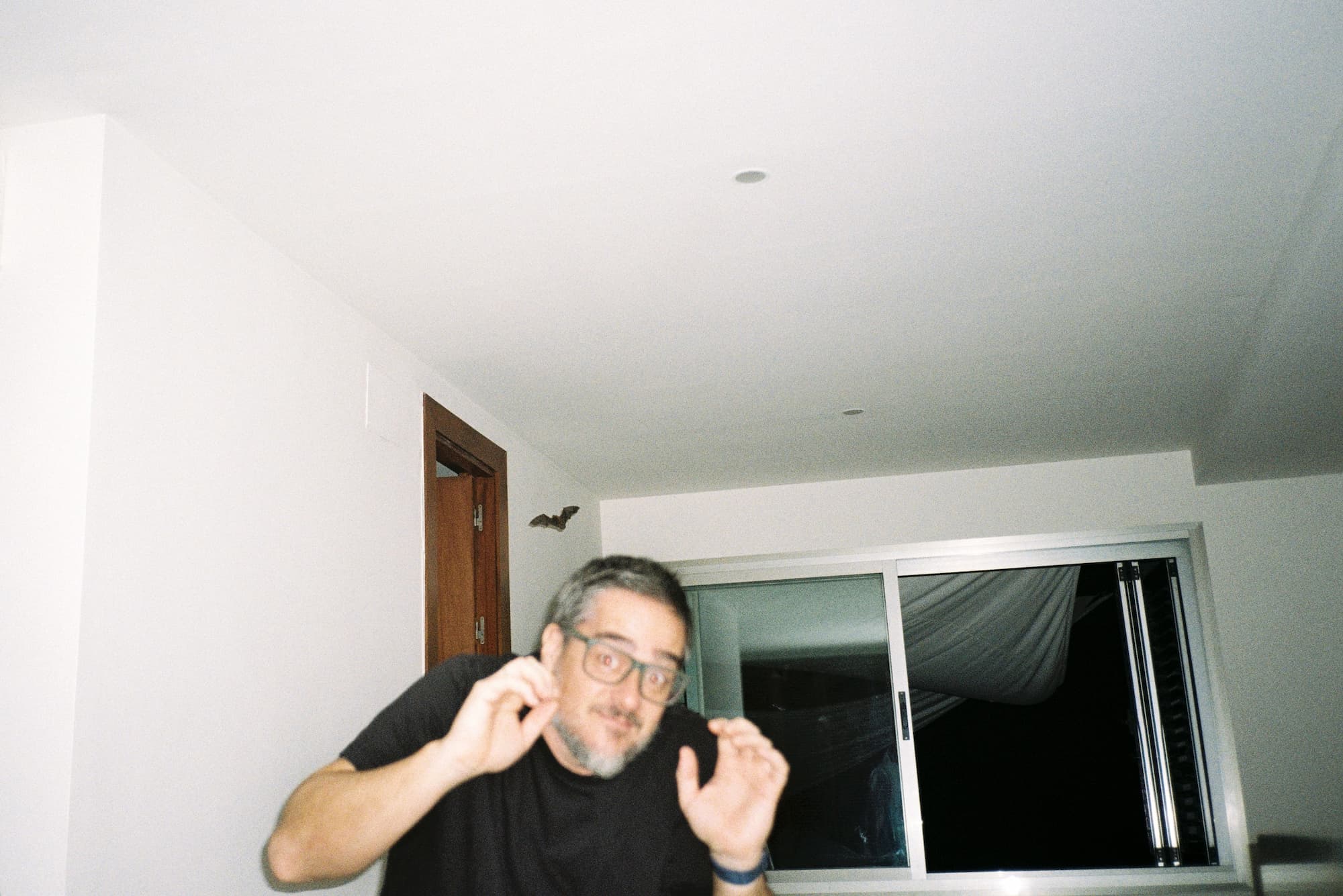 Man with glasses appears startled inside a white room while a bat flies near the ceiling above an open window and wooden door.