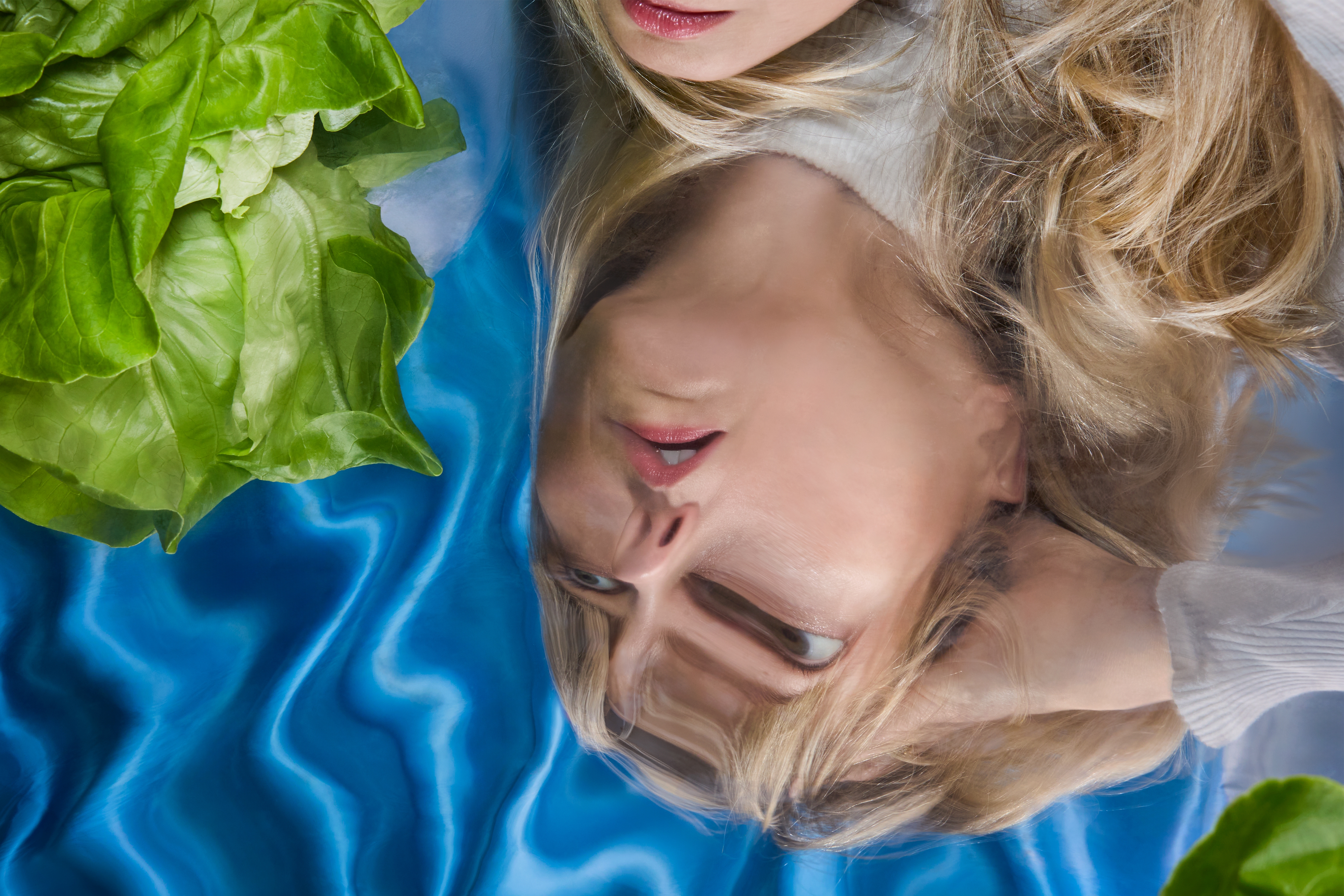 A woman’s face appears distorted in the reflection of a rippling, colorful tabletop next to a head of fresh butter lettuce.