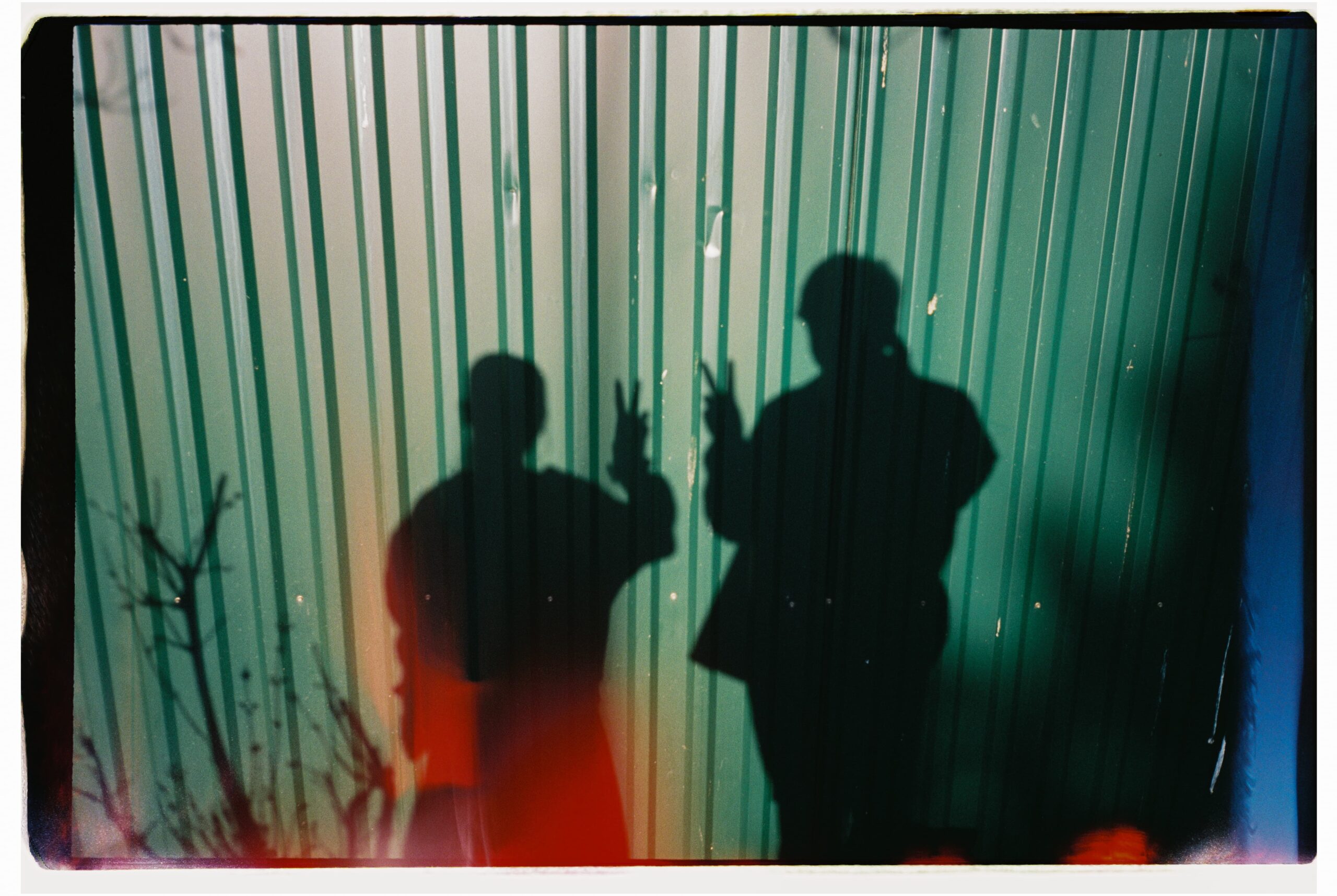 Two People Shadows Showing Peace Signs on Metal Wall Film photograph of two people shadows making peace signs on a textured metal wall, with a visible film light leak.
