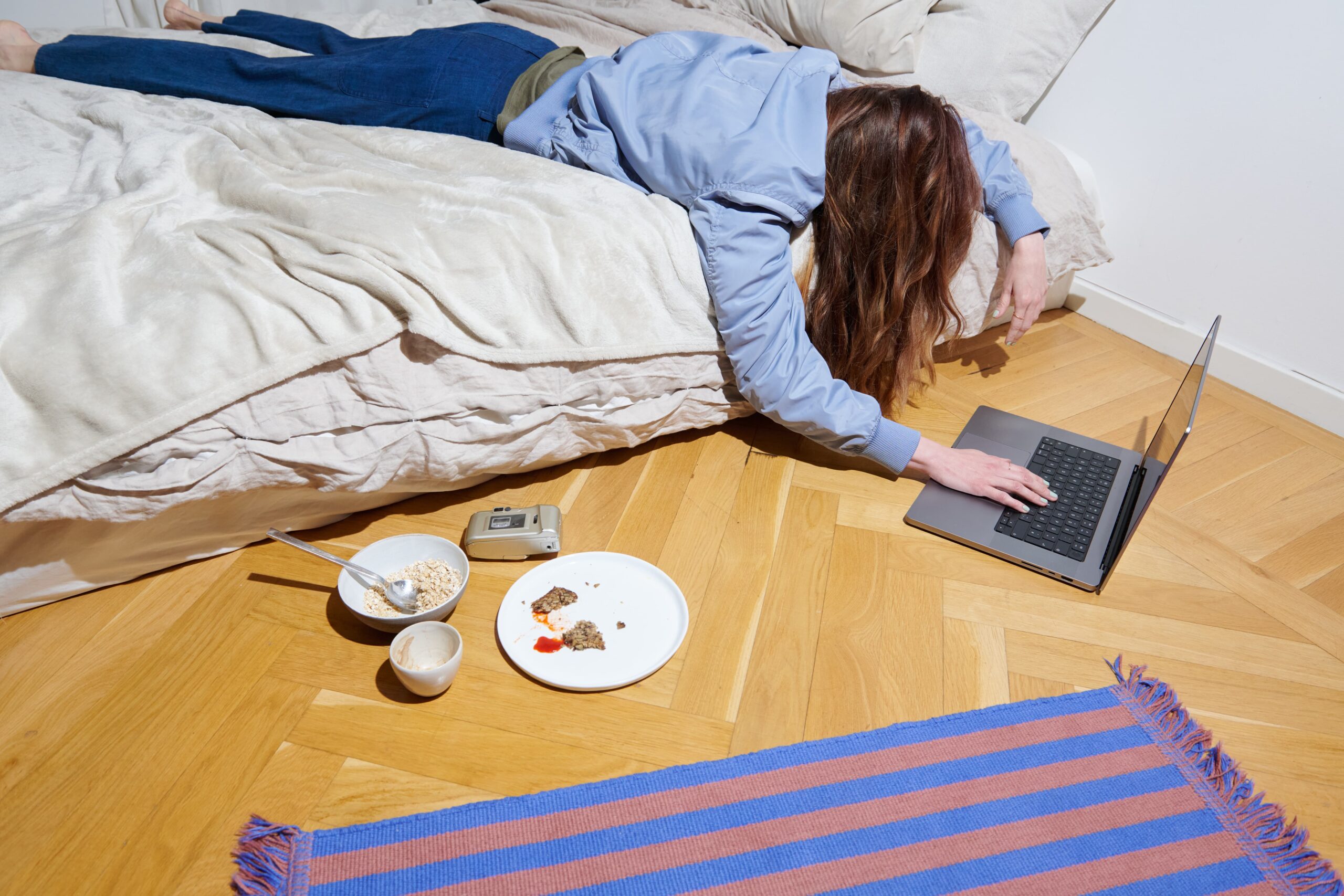 Between Working and Sleeping A person lies across a bed with one arm reaching down to a laptop on the floor. Nearby, a plate with partially eaten food, a bowl, and a small analogue camera are placed on the parquet floor.